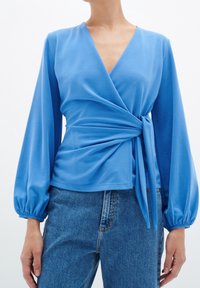 Blå omslagsblus med långa, puffiga ärmar och knytning i midjan, tillverkad av mjukt tyg. Har en djup V-ringning och slät textur.