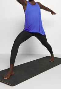 Top de tirantes morado combinado con leggings negros, ambos hechos de material elástico. Persona en una postura de yoga sobre una esterilla de ejercicio oscura.