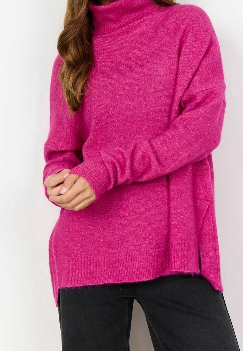 Pull à col roulé fuchsia avec une coupe ample, des poignets côtelés et des fentes latérales ; texture douce qui complète un pantalon noir.