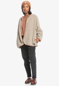 Ljus beige, texturerad fleecejacka med långa ärmar, kontrasterande brun trim och sidofickor. Matchad med en brun topp, svarta jeans och bruna skor.
