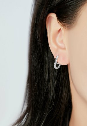 Pendiente de aro de plata con un diseño retorcido, que presenta una superficie texturizada y un pequeño gancho. Mostrado en la oreja de un modelo con cabello oscuro.