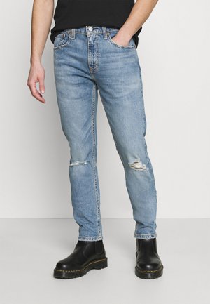 Personne portant un jean bleu clair déchiré, des bottes noires et une chemise noire, debout avec une main dans la poche devant un fond uni.