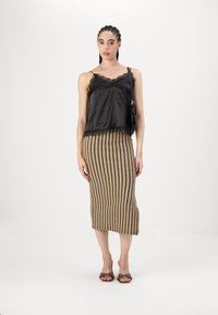 Melna satīna kamzola ar mežģīņu apdari, saskaņota ar adītu beige svārku, kuram ir viļņveidīga svītru raksts. Modelis valkā brūnas papēžsandales.