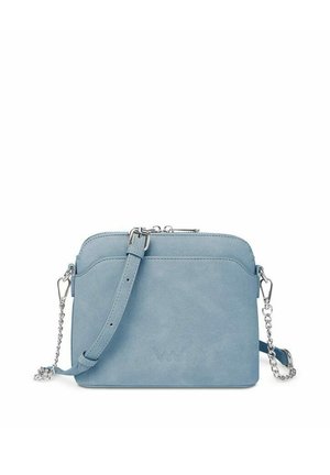 Woman in white coat and knit hat stands by snow-covered pine tree holding a small blue purse, looking back and smiling.