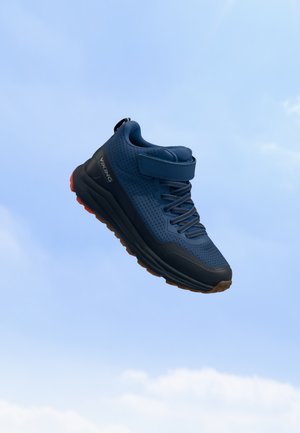 Baskets bleues et noires avec bride en velcro et lacets, semelle noire avec bande de roulement marron, montrées flottant sur un fond de ciel bleu clair.