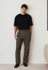 Schwarzes kurzärmeliges Hemd kombiniert mit braunen gefalteten Hosen und schwarzen Schuhen. Das Outfit zeichnet sich durch eine lockere Passform und ein einfaches Design aus.