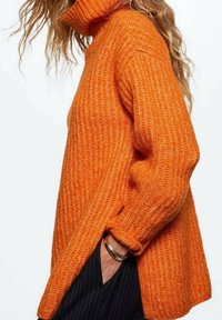 Personne portant un pull à col roulé en maille orange surdimensionné avec une texture côtelée, main dans la poche, et de longs cheveux ondulés partiellement visibles.