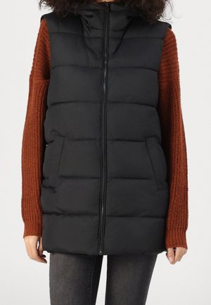 Zwarte gewatteerde mouwloze bodywarmer met rits aan de voorkant, gedragen over een roestkleurige gebreide trui en donkere spijkerbroek door een persoon staand tegen een witte achtergrond.