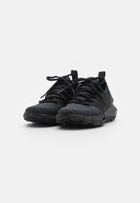 Un par de zapatillas deportivas negras con suelas texturizadas y parte superior de tela de punto, con cordones gruesos atados de forma holgada, sobre un fondo blanco liso.