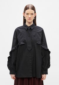 Črna srajca z nagubanimi poudarki na rokavih in prsih, z izrazitim ovratnikom in detajlom z vrvico. Narejena iz gladkega blaga, sproščen kroj.