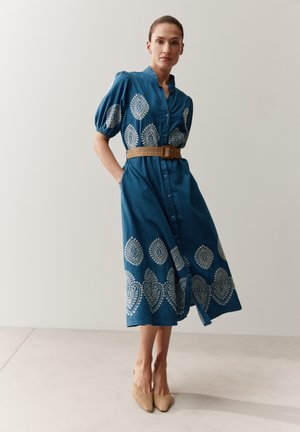Frau, die ein blaues knielanges Kleid mit weißer Stickerei, Puffärmeln, einem braunen geflochtenen Gürtel und beigen spitzen Schuhen trägt, stehend in Innenräumen.
