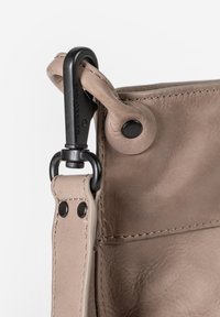 Tan leren tas met een gladde textuur, voorzien van een zwarte metalen clip en een cirkelvormige knoopaccent. Gestikte details langs de naden.