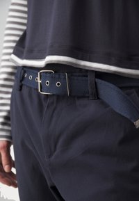 Pantalons en coton bleu marine avec un tissu texturé, dotés d'une large ceinture bleu marine avec une quincaillerie en métal, et d'un superposé en chemise rayée grise et blanche.