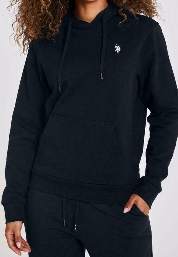 Femme portant un sweat à capuche noir avec un petit logo blanc brodé sur la poitrine et un pantalon de sport noir assorti.