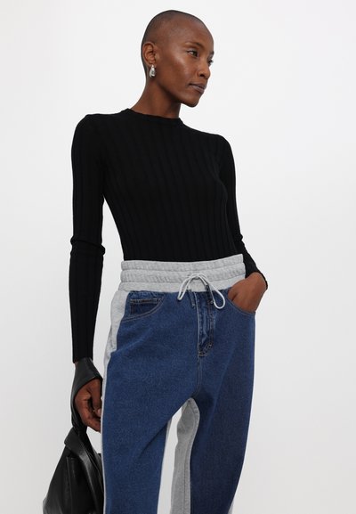 Top de manga larga de canalé negra combinado con pantalones de chándal en colores gris y azul con bloques de color, que cuentan con una cintura elástica y cordón. Bolsa negra en la mano.