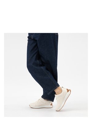 Persona che indossa jeans blu scuro e sneakers bianche con suola marrone, in piedi con un piede leggermente sollevato contro uno sfondo bianco.