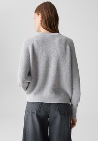 Femme aux cheveux bruns portant un pull en tricot gris clair et un jean ample gris foncé, photographiée de dos contre un fond uni.