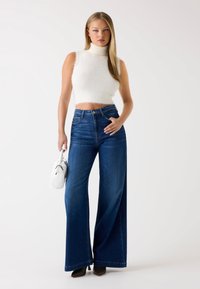 Suéter de tortuga corto y esponjoso de color blanco con jeans de mezclilla azul de tiro alto y pierna ancha. La modelo sostiene un pequeño bolso blanco y lleva tacones negros.