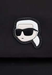 Borsa in tessuto nero con una spilla a forma di faccia di cartone animato bianca e nera con occhiali da sole e acconciatura intrecciata. Texture liscia con dettagli cuciti.