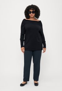 Pulover negru, cu umerii descoperiți și detalii cu dungi, asortat cu pantaloni indigo. Modelul poartă ochelari de soare negri și pantofi negri cu talpă plată.