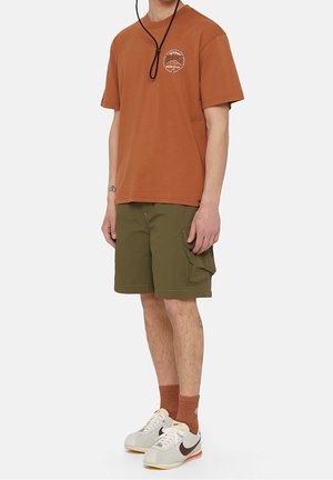 T-shirt en coton marron avec un logo, shorts cargo vert olive et baskets blanches avec des accents rouges et beiges. Porté avec des chaussettes orange.
