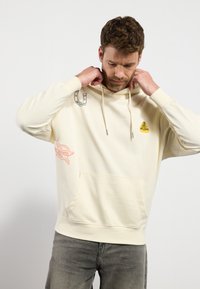 Cremefarvet hoodie lavet af blødt stof, med en frontlomme, snørrepullover og trykte grafik i blåt og rødt.