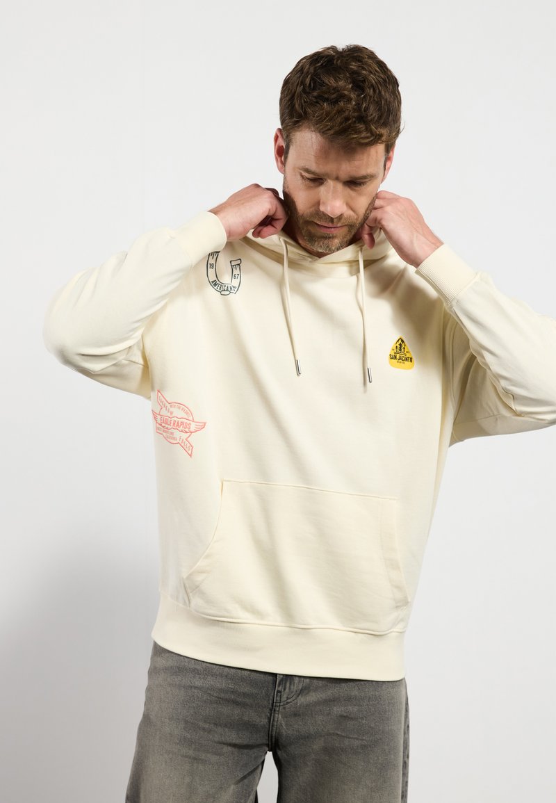 Cremefarvet hoodie lavet af blødt stof, med en frontlomme, snørrepullover og trykte grafik i blåt og rødt.