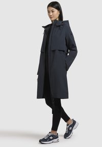 Long manteau de pluie navy avec capuche, fermeture éclair à l'avant et cordons de serrage ajustables. Porté par-dessus des leggings noirs et des baskets New Balance.