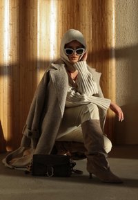 Vrouw in beige gebreide capuchon, oversized jas, zonnebril en suède kniehoge laarzen die hurkt met een zwarte handtas op de vloer tegen een houten muur.