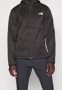 Giacca sportiva nera con chiusura a zip, tasche laterali e cappuccio. Texture liscia con logo bianco a contrasto sul petto sinistro.