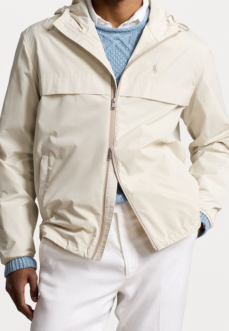 Beige lättviktsjacka med huva, dragkedja och två sidofickor. Har en bröstficka och en logotyp på vänster sida.