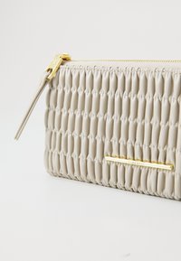Lys beige skinnclutch med en teksturerad, kviltad yta. Har en guldfärgad dragkedja och en horisontell guldaccent på framsidan.