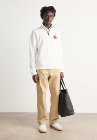 White zip-up sweatshirt with red logo, beige trousers, and white sneakers with tan accents. Black tote bag in hand. Neutral background.