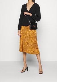 Blouse noire à manches longues avec un décolleté en V, associée à une jupe jaune mi-longue avec un motif ondulé. Sac à main noir texturé et talons noirs.