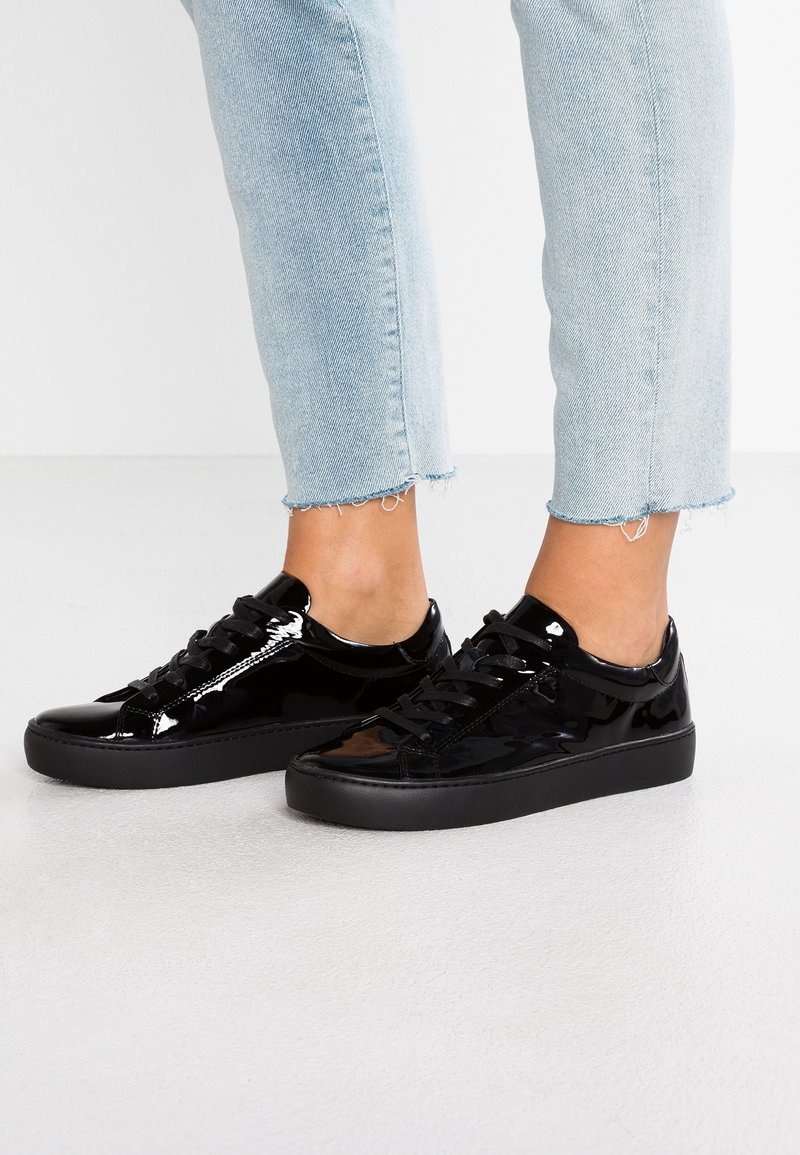 Baskets en cuir verni noir brillant avec lacets portées avec un jean court bleu clair sur un fond clair uni.