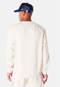 Sudadera color crema con mangas largas y puños de canalé. Presenta una textura suave y un corte relajado. Llevada con una gorra azul oscuro.