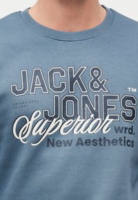 Μπλε φούτερ με το κείμενο «JACK & JONES Superior New Aesthetics» τυπωμένο μπροστά με έντονα και πλάγια γράμματα.