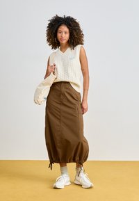 Suéter sin mangas de punto beige, falda larga marrón con dobladillo ajustable, llevando un bolso beige, zapatillas blancas. Tejido texturizado, corte relajado.