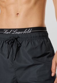 Torso masculino vestindo calções pretos com cordão de ajuste e uma cintura preta visível rotulada "Karl Lagerfeld" em escritura branca.