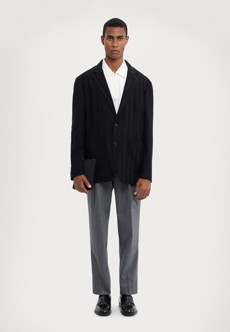 Blazer à chevrons noir avec deux poches avant, porté sur une chemise blanche, associé à un pantalon gris et des chaussures noires, tenant un portefeuille à la main.