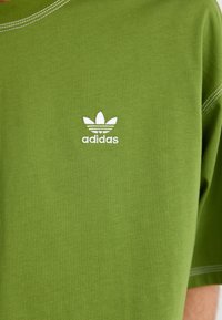 T-shirt en coton vert avec col ras du cou, présentant le logo blanc d'Adidas et l'emblème à trois bandes sur la poitrine. Texture lisse, coupe décontractée.