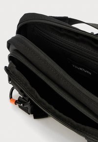 Μαύρη υφασμάτινη τσάντα με πολλαπλές θήκες, λεία φερμουάρ, υφασμάτινες λεπτομέρειες και ένα μικρό πορτοκαλί κλιπ προσαρτημένο για επιπλέον λειτουργικότητα.