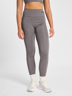 Vrouw met grijze leggings met hoge taille, witte sokken en witte sportschoenen, staand tegen een effen lichte achtergrond.
