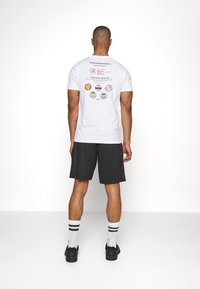 White t-shirt displaying various baseball logos and text on the back. Paired with black mesh shorts, white socks with black stripes, and black shoes.