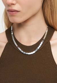 Metalhalskæde med kvadratiske led, skinnende sølvfinish, båret over en brun ribbet bluse. Designet har et struktureret og geometrisk mønster.