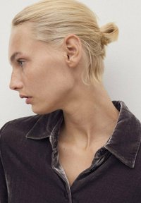 Perfil de una persona con cabello rubio recogido en un moño bajo, que lleva una camisa de terciopelo gris oscuro con una textura suave y cuello puntiagudo.