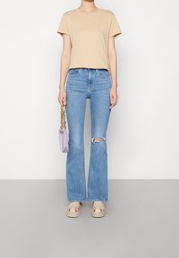 T-shirt beige à manches courtes, jean évasé bleu clair avec une déchirure au genou, sandales crème, et petit sac à main violet avec une bandoulière chaîne dorée.