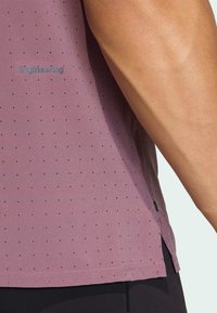 Αθλητική μπλούζα με κοντά μανίκια σε λιλά χρώμα, με διάτρητο σχέδιο και ανοίγματα στα πλάγια, με λογότυπο σε ανοιχτό μπλε στην επάνω αριστερή πλευρά.