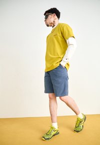 Gelbes Baumwoll-T-Shirt, übereinander getragen mit einem weißen Langarmshirt, kombiniert mit blauen Cargo-Shorts und grün-gelben Sneakers.