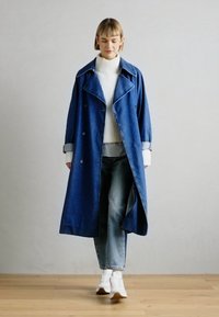 Trench azul de mezclilla con grandes solapas, botones de doble botonadura y puños enrollados, usado sobre un cuello de tortuga blanco de canalé y vaqueros.